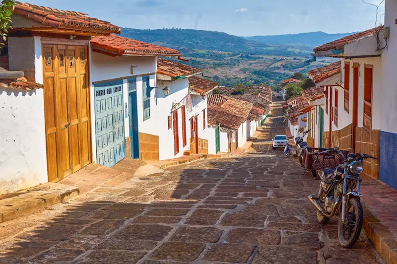 Cobblestoned street sloping downhill between colourful red-tiled houses and a view of countryside beyond in Barichara, Colombia.