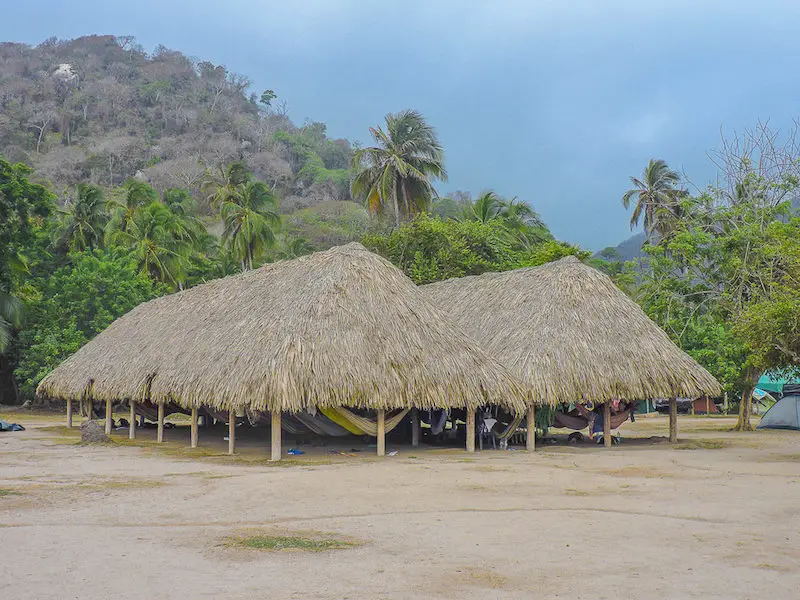 Rows of hammocks under a palapa roof on the beach backed by palm trees in Tayrona National Park, Colombia.